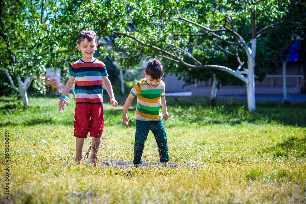 Fototapeta premium Little boy and his brother play in summer park. Children with colorful clothes jump in puddle and mud in the garden.