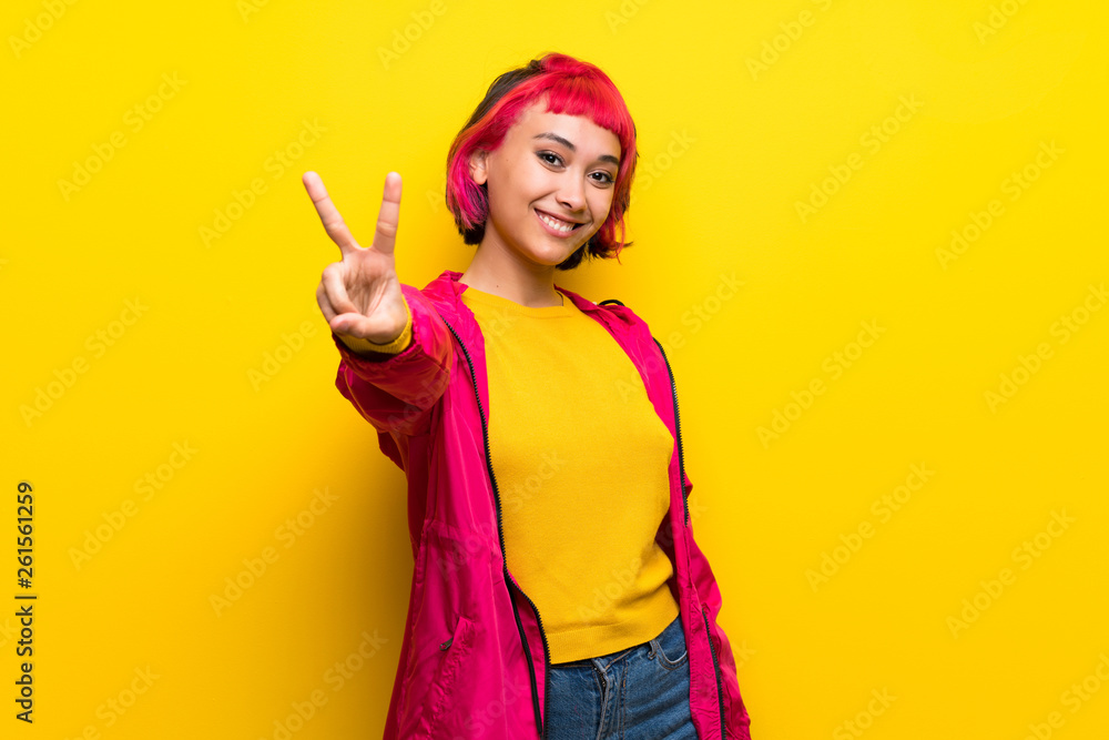 Young woman with pink hair over yellow wall smiling and showing victory sign