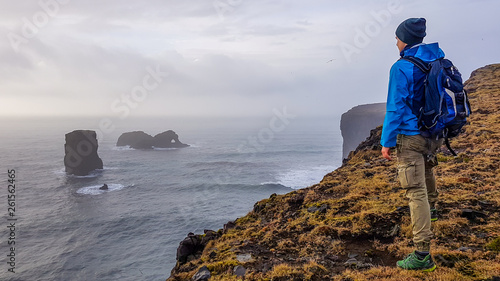 A young man wearing blue jacket, standing at the edge of a steep cliff. Some rock formations poking out of the sea. Moody atmosphere. Pure happiness and enjoyment derived from traveling photo