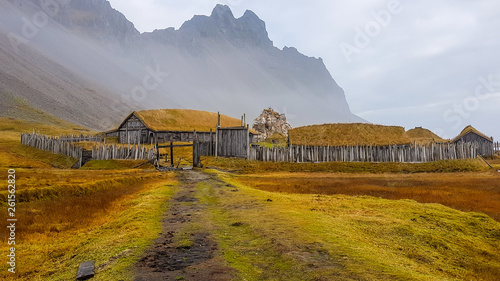 An abandoned vikings village. Sod rooftops, turf rooftops. Village located at the bottom of a high mountain. Around the farm a wooden fence. Dry grass all around. Traditional Scandinavian architecture photo