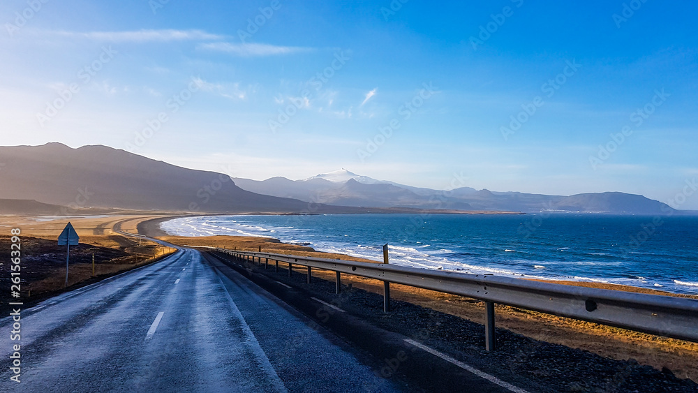 An endless road along the coast. Road stretches over the horizon. Waves gently wash the shore. Empty road, with no cars pasing by. In the back tall mountains emerge from the seashore. Road trip.