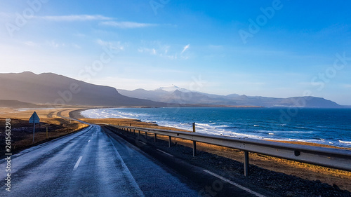 Obraz na płótnie An endless road along the coast. Road stretches over the horizon. Waves gently wash the shore. Empty road, with no cars pasing by. In the back tall mountains emerge from the seashore. Road trip. w salonie