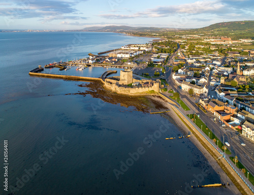 Belfast Lough. Medieval Norman Castle in Carrickfergus in sunrise light. Aerial view with marina, yachts, parking, breakwater, groin, sediments and far view of Belfast in the background