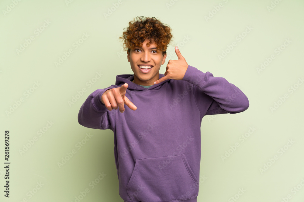 Young african american man over isolated green wall making phone gesture and pointing front Young african american man over isolated green wall making phone gesture and pointing front