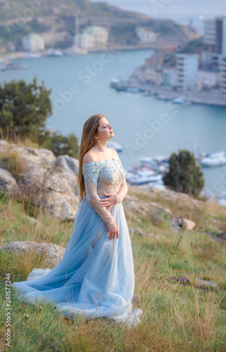 A girl with long hair on the beach. She's wearing a beautiful blue dress . In the background view of the Bay, sea and city. Soft focus.