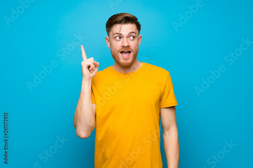 Redhead man over blue wall intending to realizes the solution while lifting a finger up