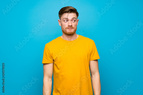Redhead man over blue wall making doubts gesture looking side