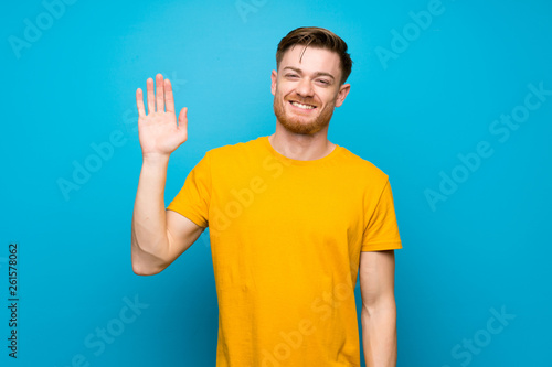 Redhead man over blue wall saluting with hand with happy expression © luismolinero