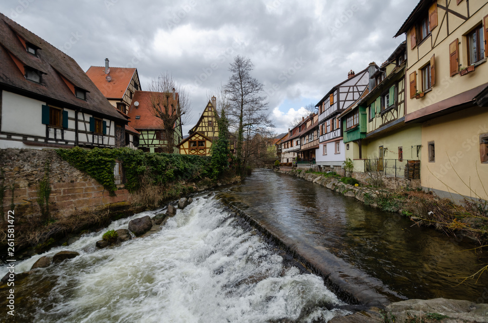 A look along the turbulent river that flows through the old town. There are old half-timbered houses with tiled roofs on both banks of the river.  There is a threshold across the river. Alsace.