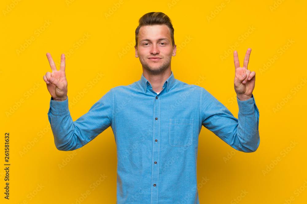 Blonde man over isolated yellow wall showing victory sign with both hands Blonde man over isolated yellow wall showing victory sign with both hands