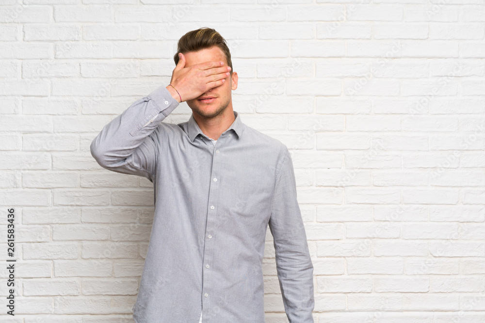 Blonde man over white brick wall covering eyes by hands. Do not want to see something Blonde man over white brick wall covering eyes by hands. Do not want to see something