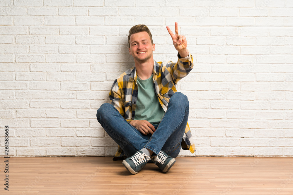 Blonde man sitting on the floor smiling and showing victory sign Blonde man sitting on the floor smiling and showing victory sign