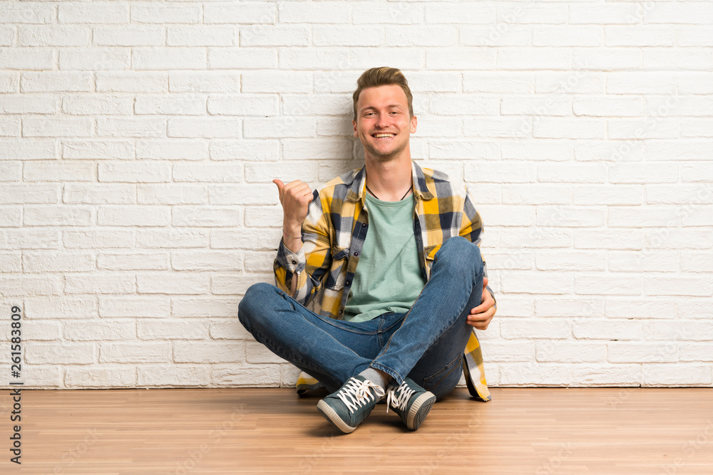Blonde man sitting on the floor pointing to the side to present a product Blonde man sitting on the floor pointing to the side to present a product