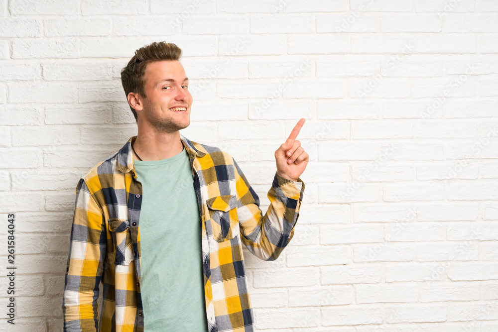 Blonde handsome man over white brick wall pointing finger to the side Blonde handsome man over white brick wall pointing finger to the side