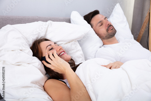 Woman Talking On Smartphone Besides Her Husband Sleeping On Bed