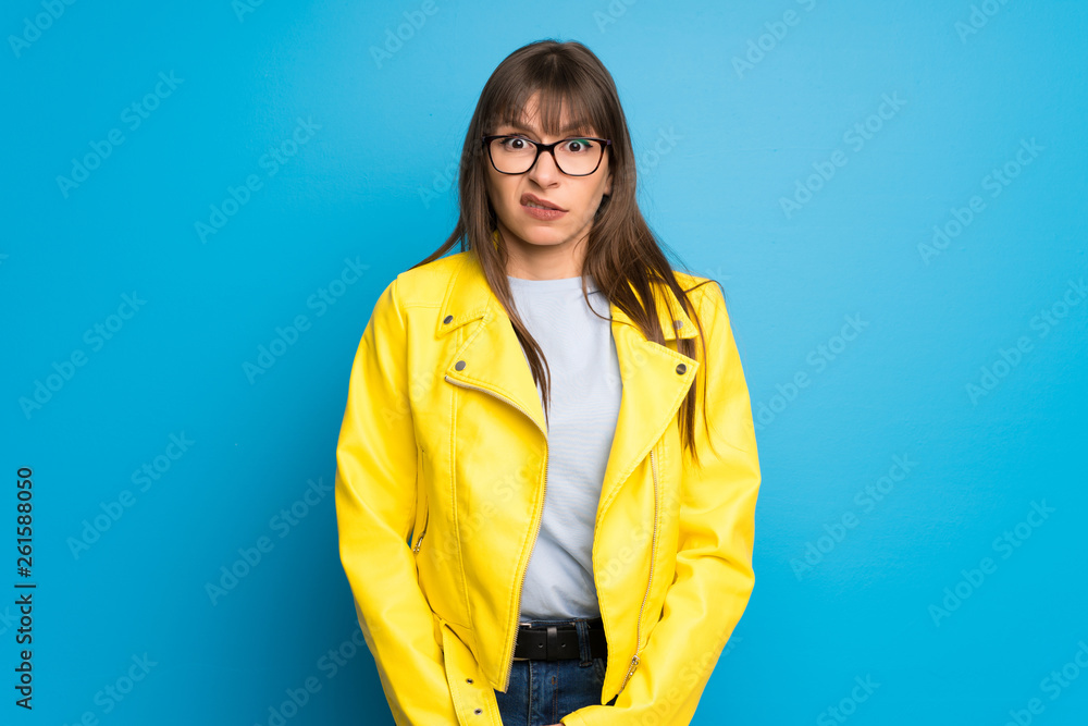Young woman with yellow jacket on blue background is a little bit nervous and scared pressing the teeth Young woman with yellow jacket on blue background is a little bit nervous and scared pressing the teeth