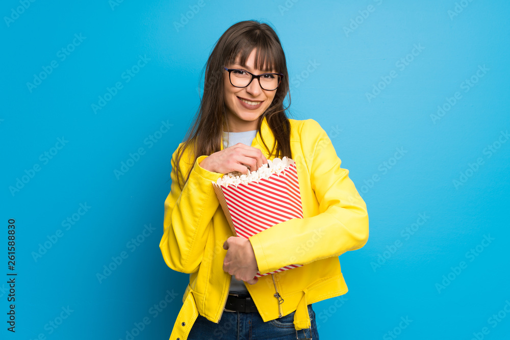 Obraz premium Young woman with yellow jacket on blue background surprised and eating popcorns