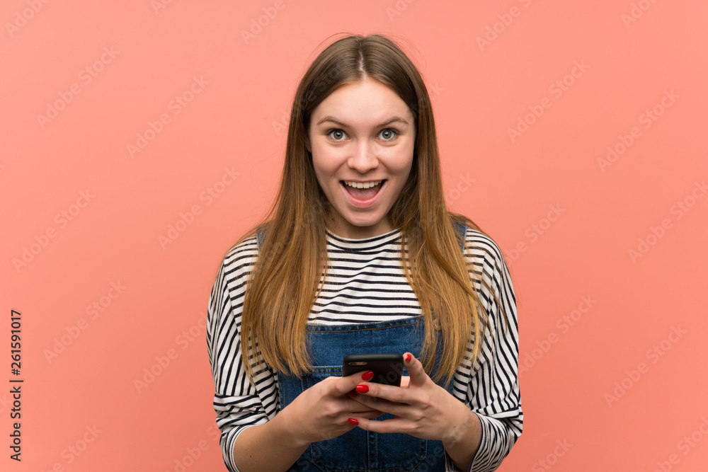 Young woman with overalls over pink wall surprised and sending a message Young woman with overalls over pink wall surprised and sending a message