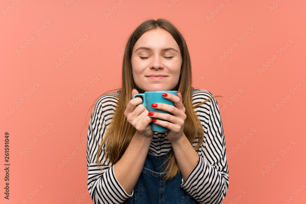 Young woman with overalls over pink wall holding a hot cup of coffee Young woman with overalls over pink wall holding a hot cup of coffee