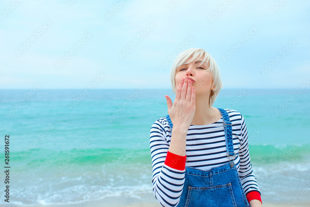 beautiful young blonde caucasian woman on vacation in striped blouse, sneakers and denim overall jumping on the beach by the amazing blue sea background beautiful young blonde caucasian woman on vacation in striped blouse, sneakers and denim overall jumping on the beach by the amazing blue sea background