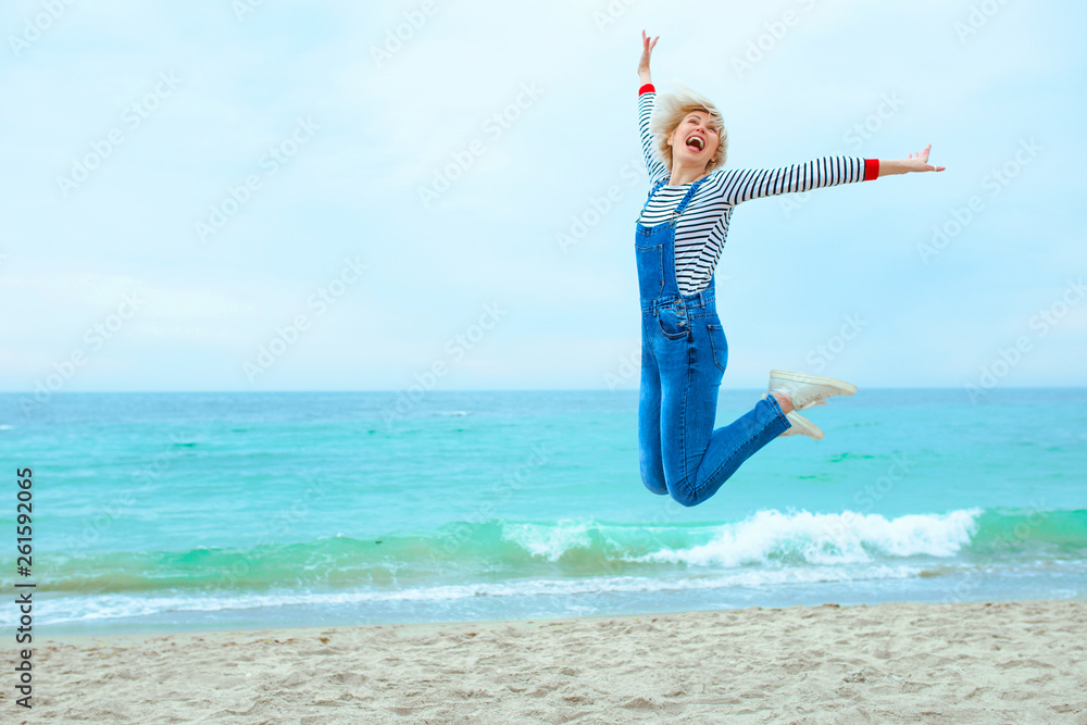 beautiful young blonde caucasian woman on vacation in striped blouse, sneakers and denim overall on the beach by the blue sea background. Happy to be at the sea beautiful young blonde caucasian woman on vacation in striped blouse, sneakers and denim overall on the beach by the blue sea background. Happy to be at the sea