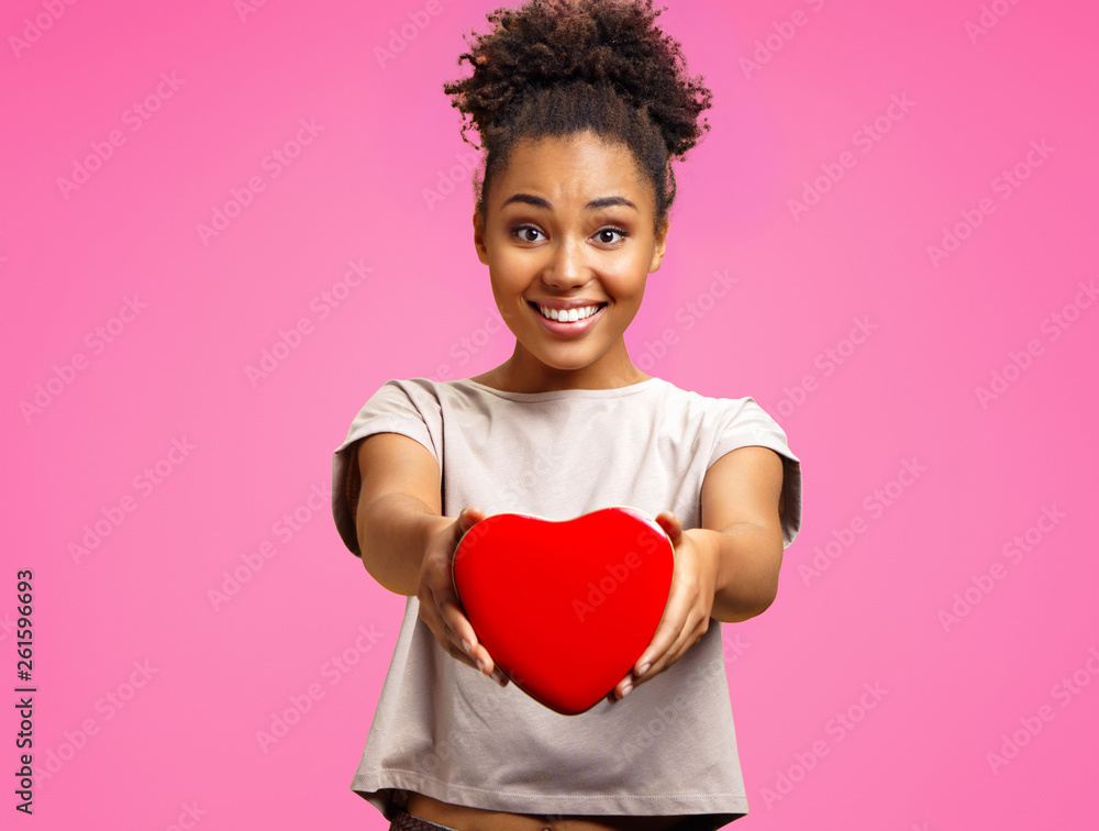 Lovely young girl holds heart shaped gift box. Photo of african american girl wears casual outfit on pink background. Emotions and pleasant feelings concept. Lovely young girl holds heart shaped gift box. Photo of african american girl wears casual outfit on pink background. Emotions and pleasant feelings concept.