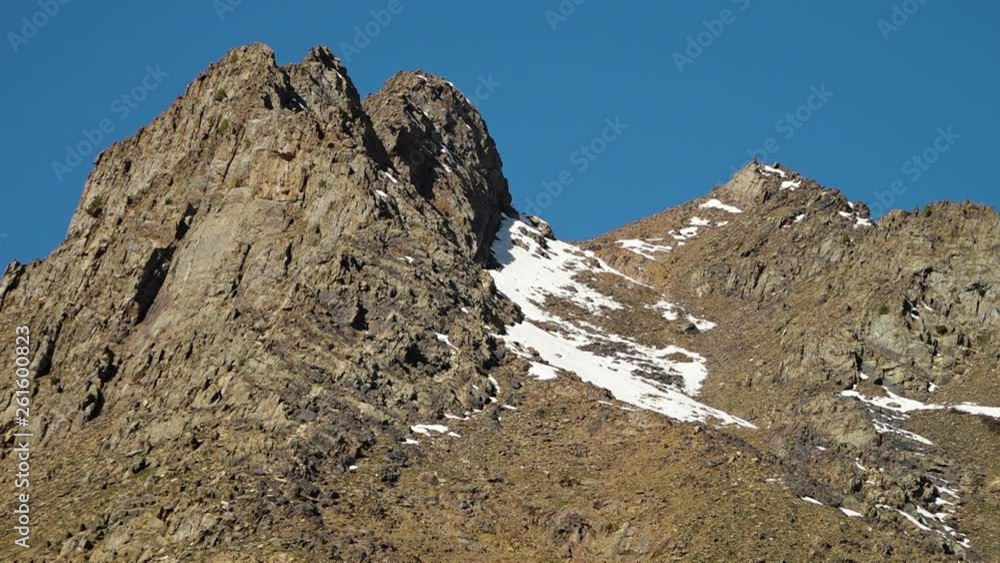 A steady, low angle, medium shot of small spots with snow over a very high rocky mountain. A steady, low angle, medium shot of small spots with snow over a very high rocky mountain.