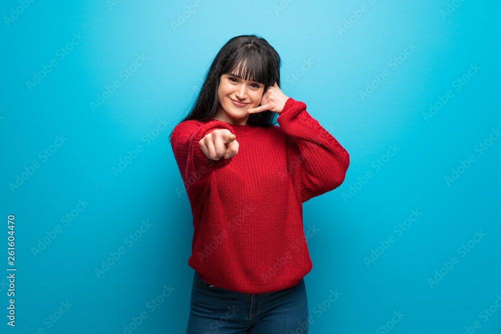 Woman with red sweater over blue wall making phone gesture and pointing front Woman with red sweater over blue wall making phone gesture and pointing front