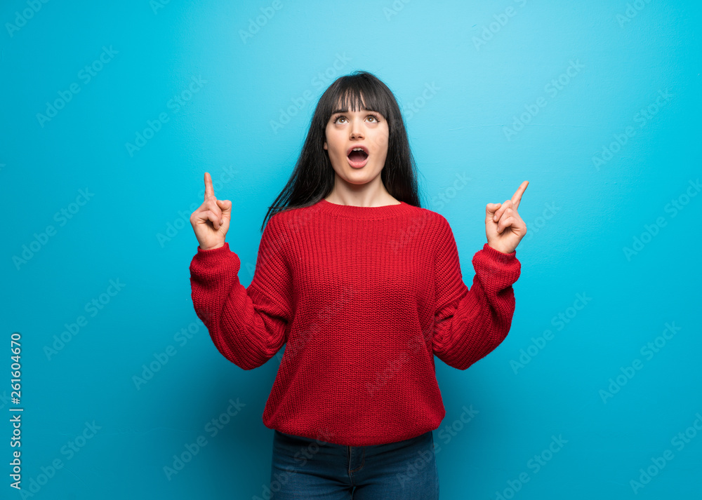 Woman with red sweater over blue wall surprised and pointing up Woman with red sweater over blue wall surprised and pointing up