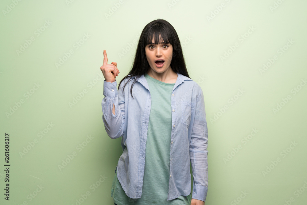Young woman over green wall thinking an idea pointing the finger up