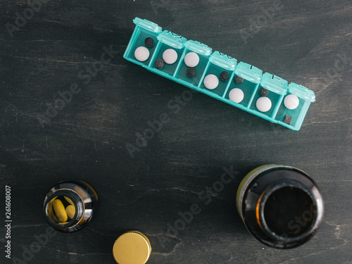 Top view of seven day pill box with pills. Green pill-box with pills visible. Open pill box and jars on dark wooden table. Copy space. Top view or flat lay.Healthy lifestyle and medical concept
