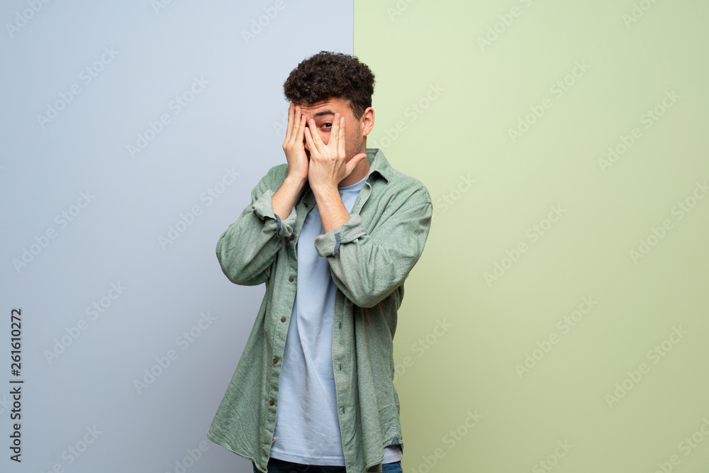 Young man over blue and green background covering eyes by hands and looking through the fingers Young man over blue and green background covering eyes by hands and looking through the fingers