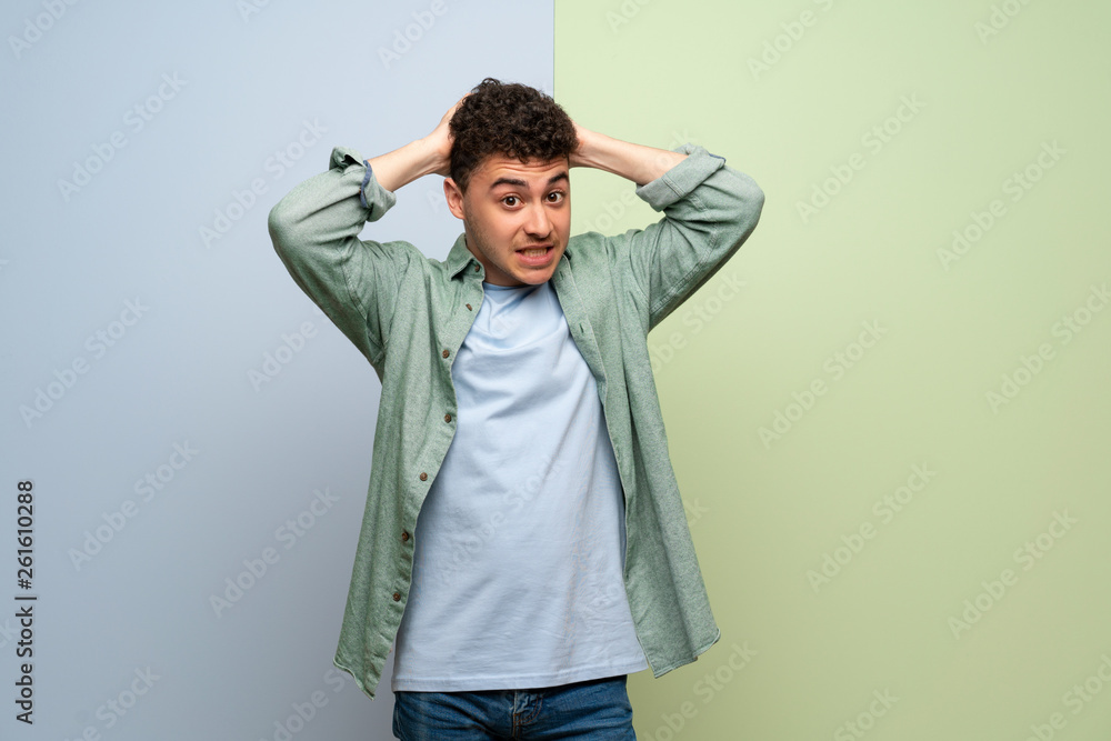 Young man over blue and green background takes hands on head because has migraine Young man over blue and green background takes hands on head because has migraine
