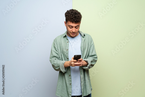 Young man over blue and green background sending a message with the mobile