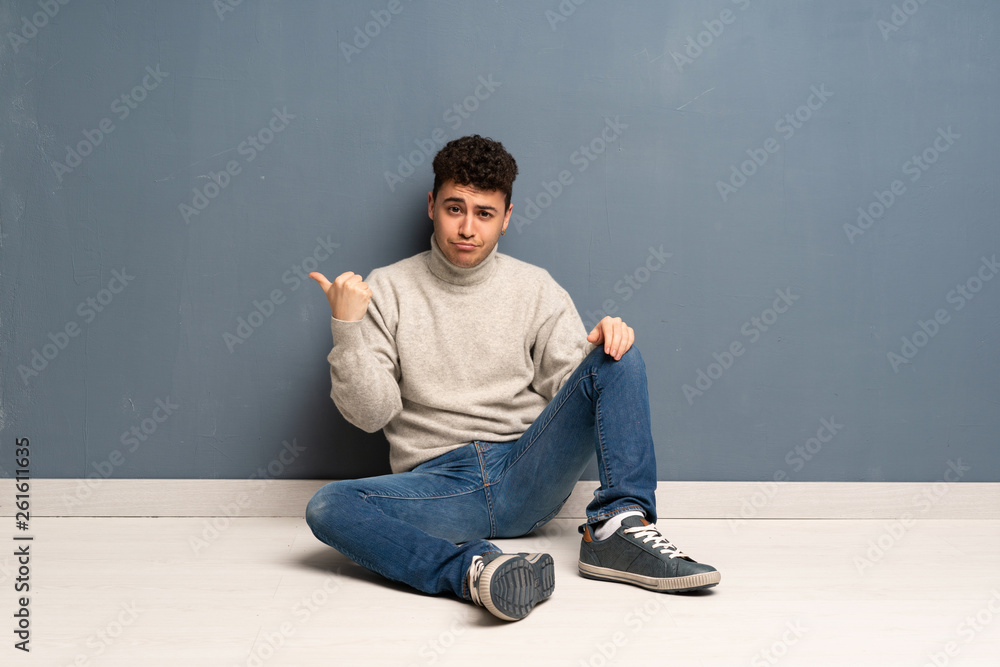 Young man sitting on the floor unhappy and pointing to the side Young man sitting on the floor unhappy and pointing to the side
