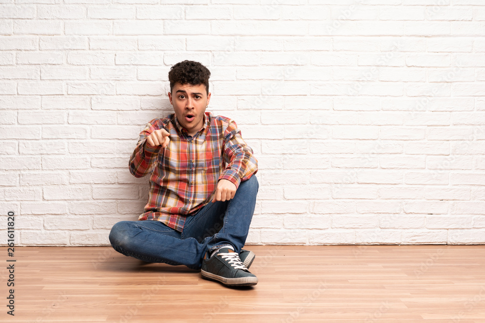 Young man sitting on the floor surprised and pointing front Young man sitting on the floor surprised and pointing front