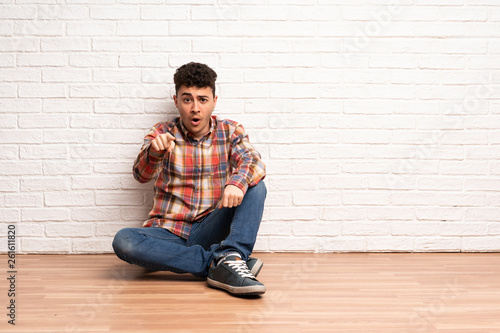 Young man sitting on the floor surprised and pointing front