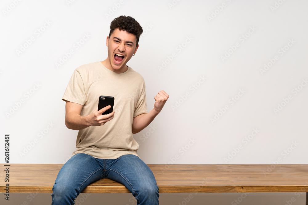 Young man sitting on table with phone in victory position Young man sitting on table with phone in victory position