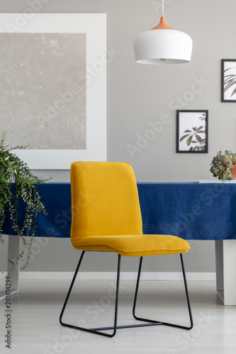 Yellow chair with metal legs placed by the table with blue tablecloth in real photo of light grey living room interior with fresh plants, lamp and posters on wall