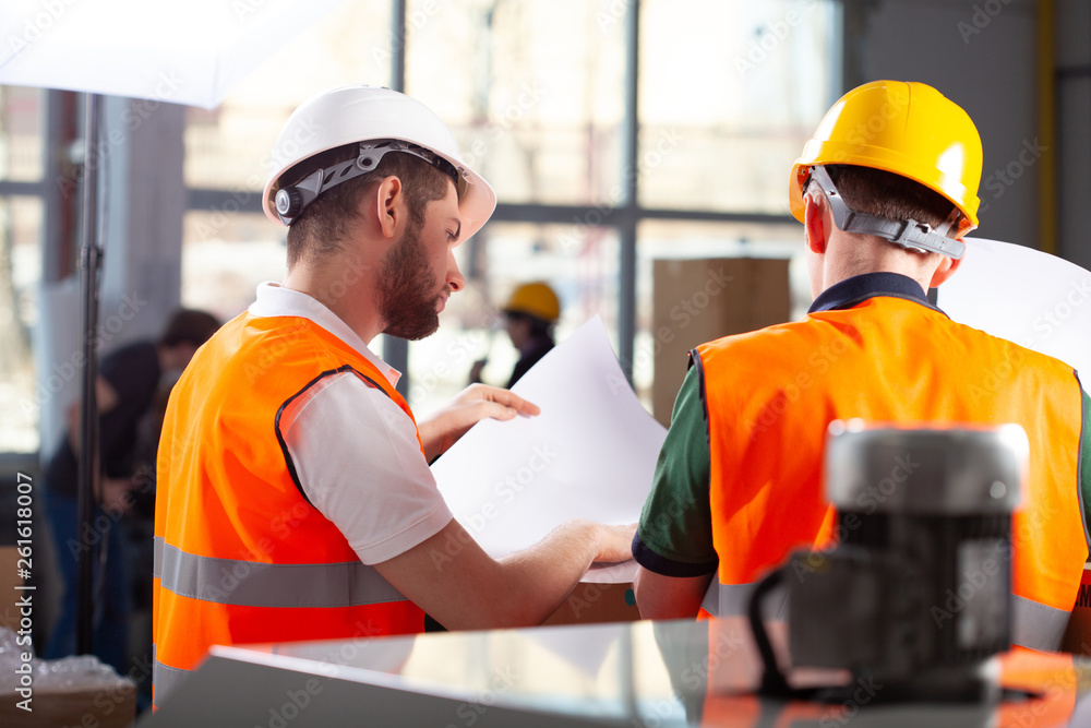 Two factory workers in orange safety jackets and helmets studying delivery documents Two factory workers in orange safety jackets and helmets studying delivery documents