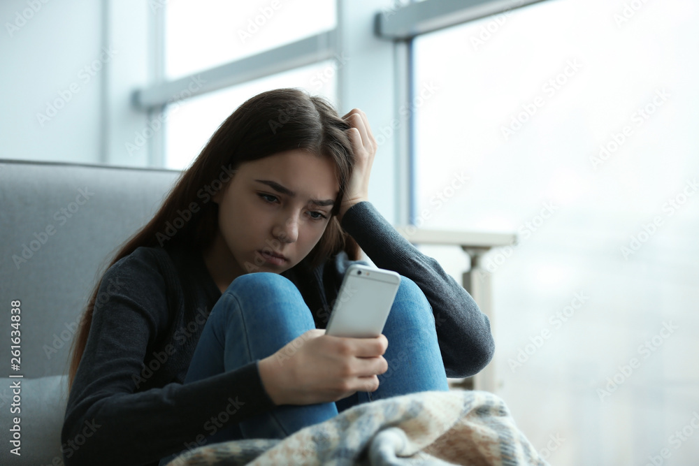 Upset teenage girl with smartphone sitting at window indoors. Space for text Upset teenage girl with smartphone sitting at window indoors. Space for text