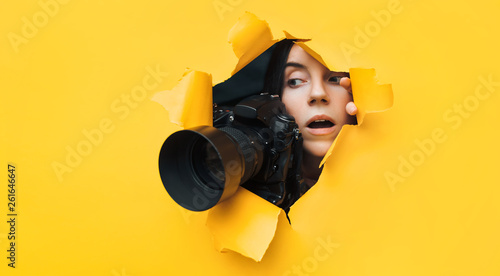 A young paparazzi girl with a reflex camera looks out from cover and looks at what is happening with her mouth open. Yellow paper, torn hole. Tabloid press. In search of the plot for photo stocks.