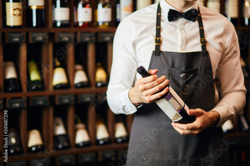 Cropped shot of wine seller or cavist in white shirt with bowtie and apron looking at bottle, telling about the origin of this sort of wine, wine shelves on the background photo
