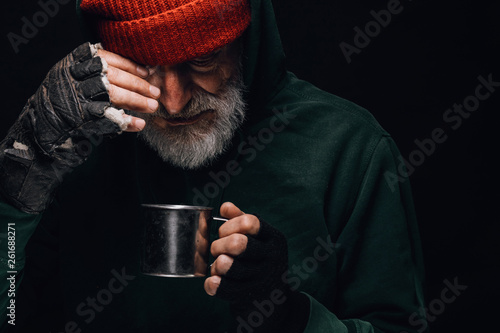 Fotografija Old homeless man with grey beard covering up in green decrepit clothes holding a