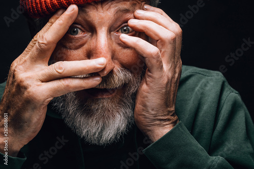 An elderly bearded man looking extremely surprised or frightened, protecting his face with hands, close up face portrait over black studio background. photo