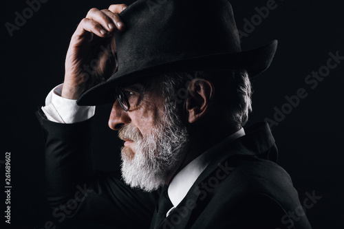 Fotografie An old male actor playing an English spy role, dressed in black suit, hiding behind fedora hat, having mysterious elegant look
