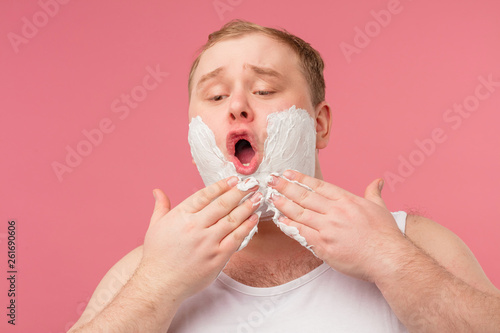 Discontent man with gel foam on cheeks  has sad expression  tired of shaving  applying foam on skin before shaving this morning feels displeased  isolated over pink background