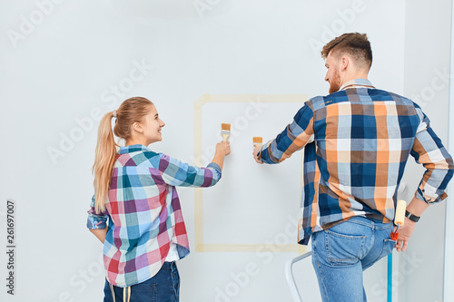 Team of two professional male and female young painters in checkered wear with brushes and rollers working indoors, making wall colour renovation of the house.