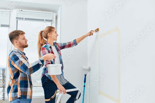 Happy girl in casual plaid shirt painting white house wall with brush and her boyfriend helps her, holding the paint bucket near her hand