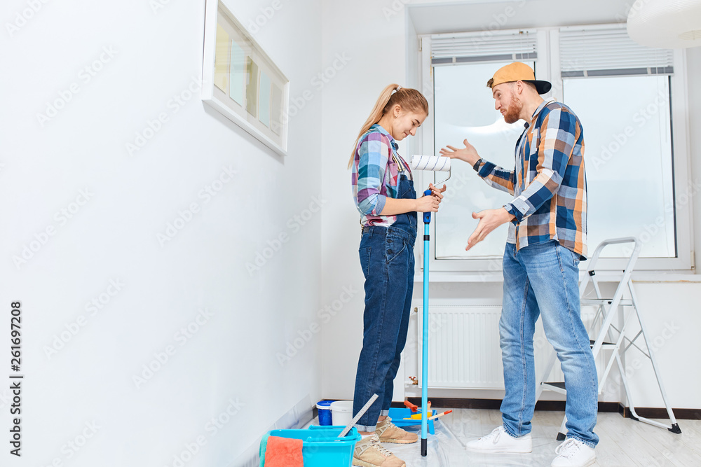Fototapeta premium Newlyweds quarrelling because of renovation in new apartment. Pissed off irritated young man scolding his embarrassed guilty wife standing in white room with set of paint tools